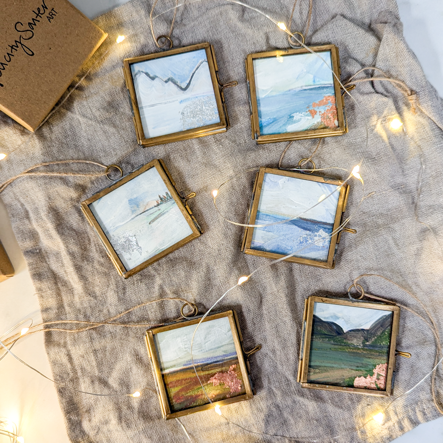 A set of six hand-painted Christmas tree decorations, each framed in a brass frame with glass windows and hanging from twine. The decorations are displayed on a fabric surface with fairy lights.